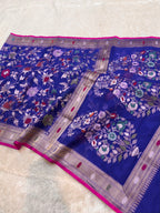 A royal blue Banarasi silk saree with intricate floral zari embroidery and a vibrant pink border, laid flat on a white textured surface.