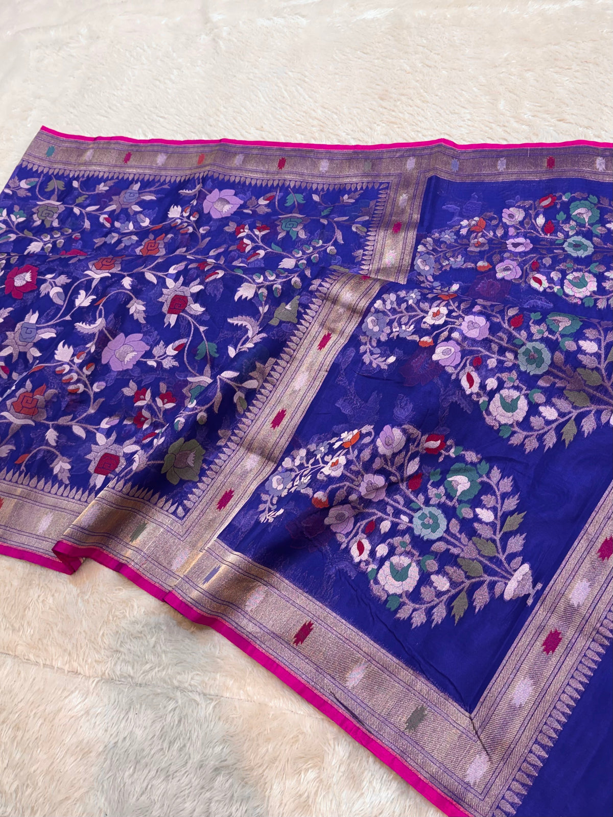 A royal blue Banarasi silk saree with intricate floral zari embroidery and a vibrant pink border, laid flat on a white textured surface.