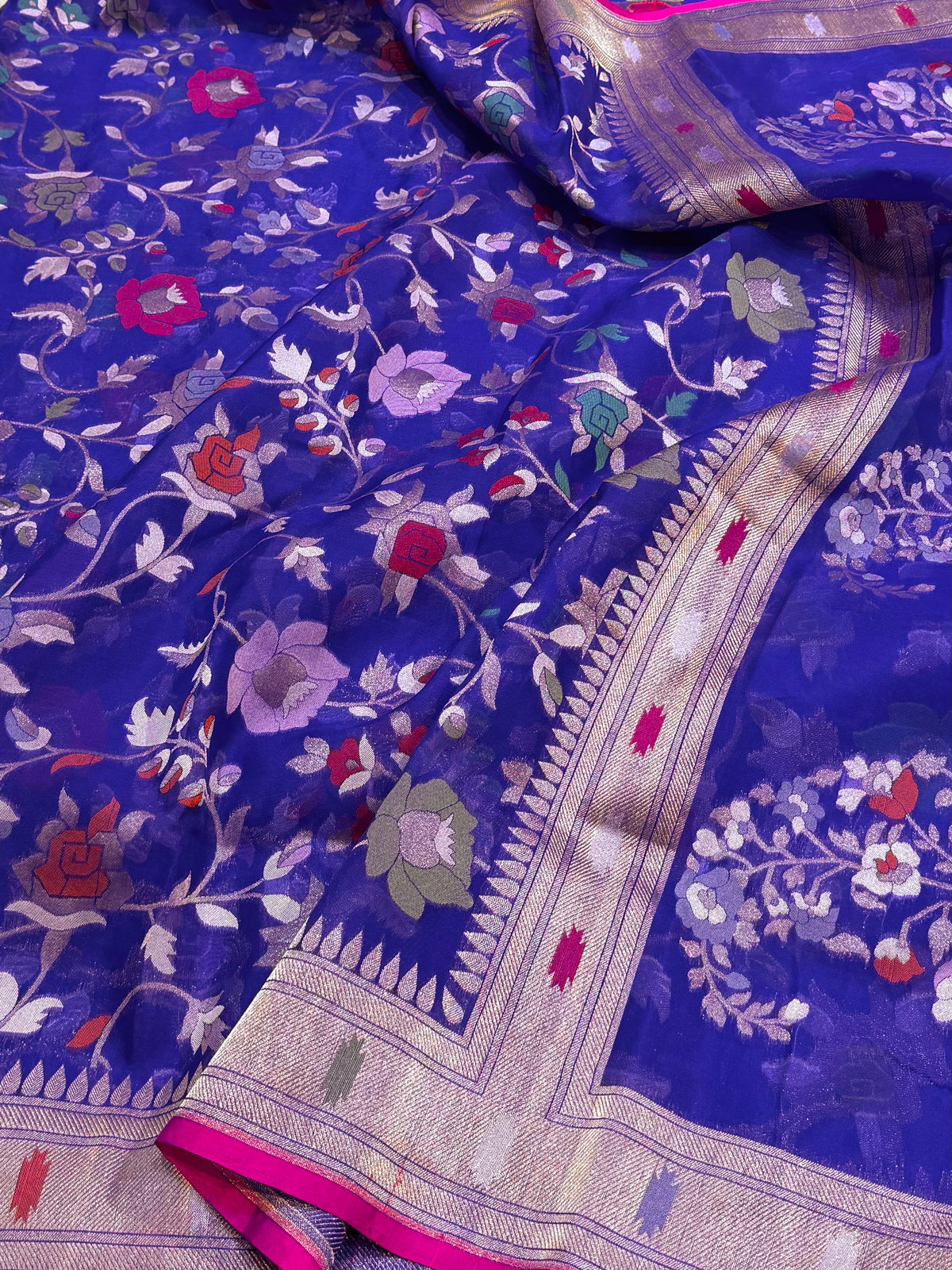 A close-up of a royal blue Banarasi silk saree with intricate floral and vine motifs woven across the fabric, featuring a detailed gold and silver zari border and a vibrant pink edge.