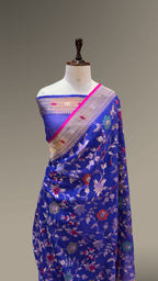 A royal blue floral silk saree draped on a white mannequin torso, featuring a pink border and a gold-toned zari border with small red and pink floral motifs.
